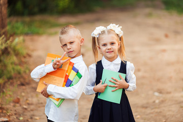 cheerful blonde babies first-graders in school uniform stand on the street with books and notebooks, the girl has white bows, children laugh