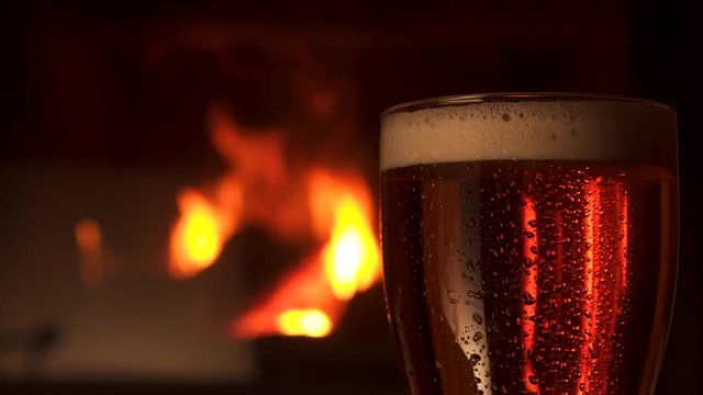 Close-up transparent glass bocal with water drops of refreshment cooled beer with foam fireplace in background. Big mug of fresh craft alcohol beverage at evening cosiness atmosphere