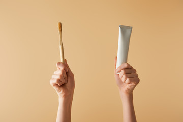 cropped view of woman holding bamboo brown toothbrush and toothpaste in tube on beige background