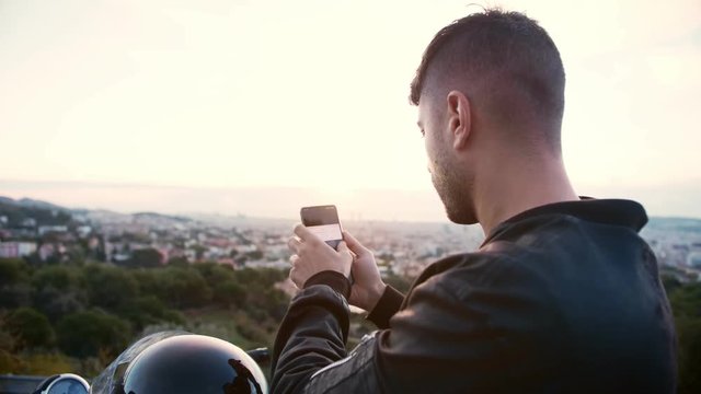 Cool and popular biker is taking a picture or photo by his smartphone or mobile phone of destination goal point beautiful stunning view of cityscape and sunrise during his road trip journey as hobby