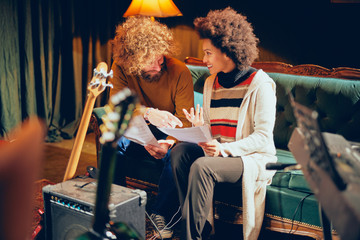 Mixed race woman sitting on the sofa with her Caucasian friend and looking at notes in home studio.
