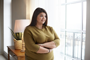 Cheerful young plus size overweight woman with black hair and chubby cheeks crossing arms on her...