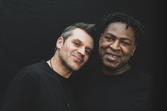 An interracial gay couple pose before the camera to have their pictures taken - black man and mixed race man with stubble - on a black background - with copy space.