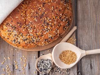 Homemade bread sprinkled with flax and sesame seeds on a wooden board under a denim fabric, next to wooden unpainted spoons with seeds