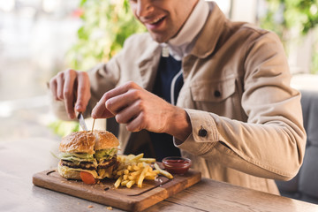 cropped view of cheerful young man cutting tasty burger on cutting board in cafe