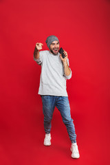 Full length photo of cheerful man 30s singing while listening to music with earphones and mobile phone, isolated over red background