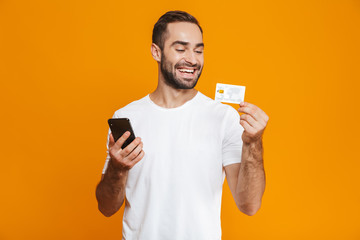 Photo of joyful man 30s in casual wear holding smartphone and credit card, isolated over yellow background