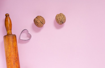 Closeup rolling pin, metal cookie cutter with shape of heart and walnuts on pink background with...