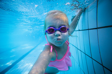 Naklejka premium Wide angle underwater photo of a toddler girl swimming in a big swimming pool with goggles and a pink bikini