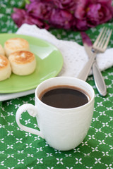 Black coffee in a white sophisticated cup, delicious dietary cheesecakes from home-made farmer cheese for breakfast, a bouquet of purple tulips on a green tablecloth.