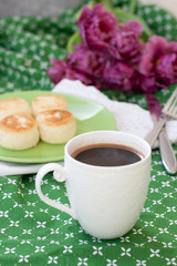 Black coffee in a white sophisticated cup, delicious dietary cheesecakes from home-made farmer cheese for breakfast, a bouquet of purple tulips on a green tablecloth.