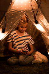 Kid in striped t-shirt using smartphone while sitting in wigwam in dark room © LIGHTFIELD STUDIOS