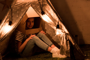 Cute kid in cardboard helmet sitting in wigwam with bulbs in dark room © LIGHTFIELD STUDIOS