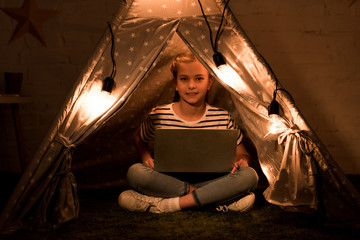 Happy kid using laptop while sitting in wigwam in dark room © LIGHTFIELD STUDIOS