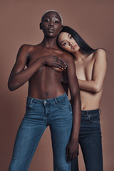 Supremely sensual. Two attractive young topless women looking at camera while standing against brown background