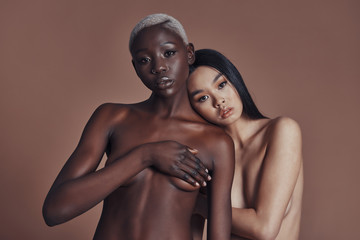 Fototapeta na wymiar Divine sensuality. Two multi-ethnic young topless women looking at camera while standing against brown background