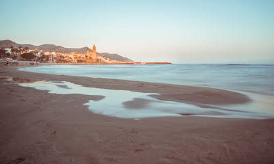Orange and teal view of mediterranean town of Sitges, with the iconic Church of Saint Bartholomew and Santa Tecla, in the suburb area of Barcelona. Blurred sea waves by a slow shutter speed