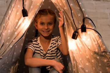 Blissful kid in striped t-shirt sitting in wigwam with hand up © LIGHTFIELD STUDIOS