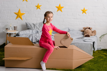 Joyful child with helmet sitting on cardboard rocket in bedroom