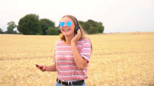 Slow motion tracking video clip of pretty blonde girl teenager young woman wearing a striped t- shirt and blue sunglasses walking listening to music on her cell phone and wireless headphones 