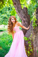 Fototapeta na wymiar A beautiful girl in a pink dress is standing by a tree