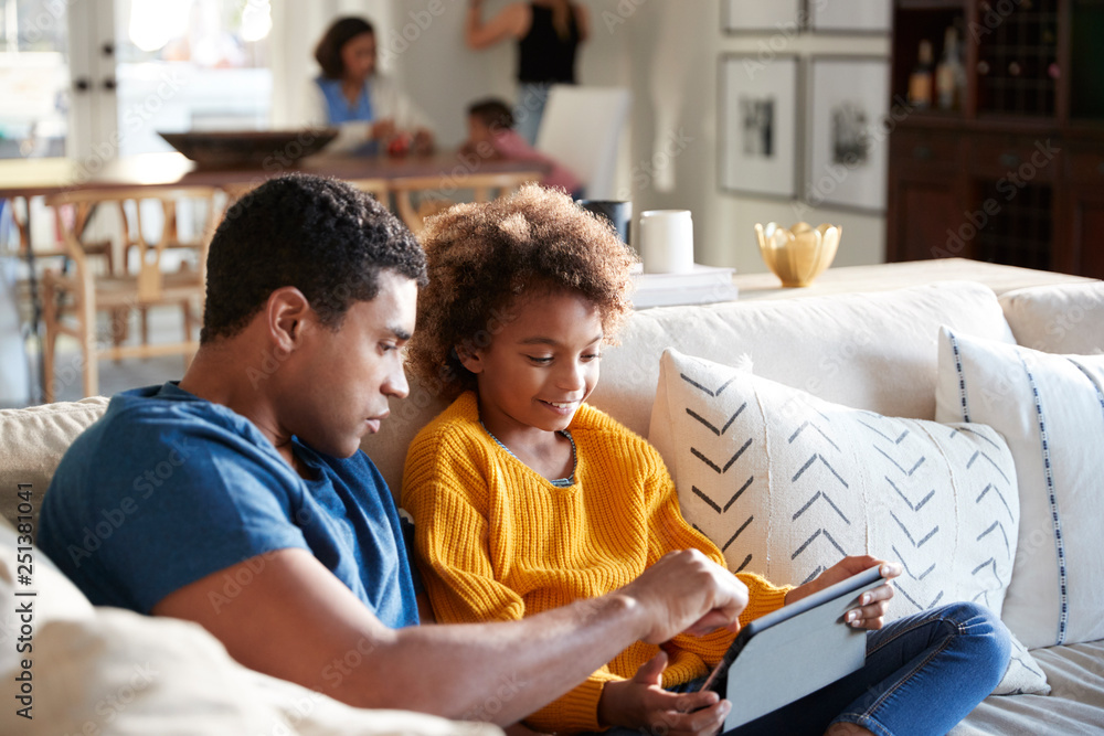 Wall mural Pre-teen girl sitting on sofa in the living room using tablet computer with her father, mother and toddler sitting at a table in the background, selective focus