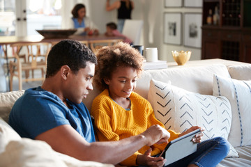 Pre-teen girl sitting on sofa in the living room using tablet computer with her father, mother and toddler sitting at a table in the background, selective focus