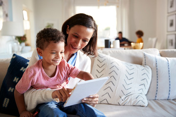 Close up of young mother sitting on a sofa in the living room with her toddler on her knee, reading him a book, father and daughter sitting at a table in the background, focus on foreground