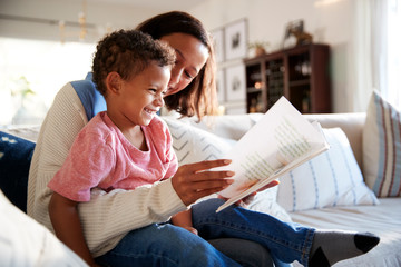 Close up of young mother sitting on a sofa in the living room reading a book with her toddler son,...
