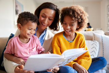 Close up of young mother sitting on sofa in the living room with her children, reading them a book,...