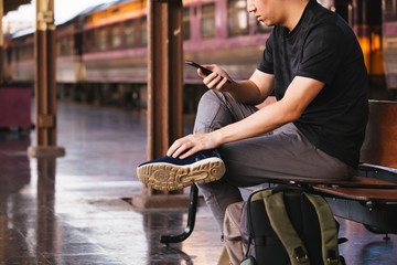 Young male tourist and backpacker using a phone while waiting for a public transportation inside train platform.