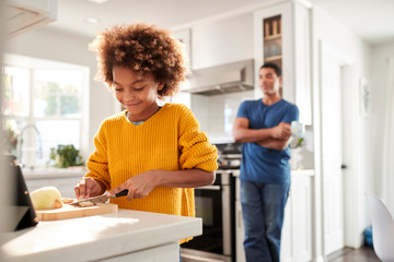 Pre-teen girl standing at worktop in the kitchen chopping, her father stands leaning in background...