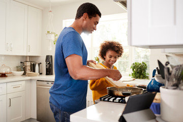 Pre-teen girl and her father standing at hob in the kitchen preparing food in a frying pan,...