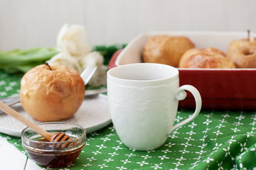 Baked apples in red, a white cup of tea or coffee, honey, a bunch of white tulips lie on a green tablecloth.