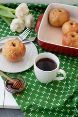 Baked apples in red, a white cup of tea or coffee, honey, a bunch of white tulips lie on a green tablecloth.