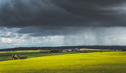 Grey rainy sky above the green meadow. Northern Ireland landscape. Dense dark clouds under the grassland and village. Stunning countryside Irish scenery. Horizon panoramic view. Contrast colors.