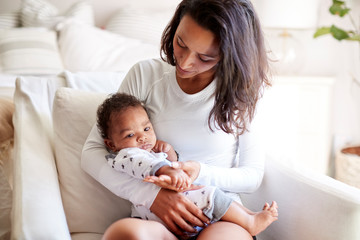 Young adult mother sitting in an armchair in her bedroom, holding her three month old baby son in...
