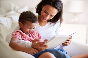 Close up of mixed race young adult mother sitting in an armchair reading a book with her three year old son on her knee, laughing, close up