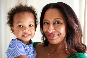 Happy mixed race middle aged grandmother holding her two year old grandson, both looking to camera...