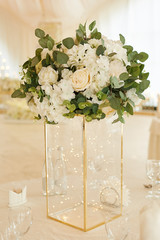 Expensive glass vase with a garland inside and a white bouquet of fresh flowers on the festive table in the banquet hall