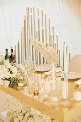A table for newlyweds of golden color decorated with white candles and flowers. Garland with white light in the background.