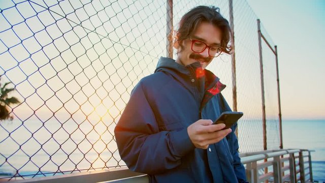 Young happy millennial hipster man skater smiling and laughing using mobile phone or smartphone application or app checking social network news feed to be up to date and being connected to internet