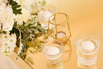 White round candles in glass candlesticks next to a bouquet of white flowers on a table in gold color