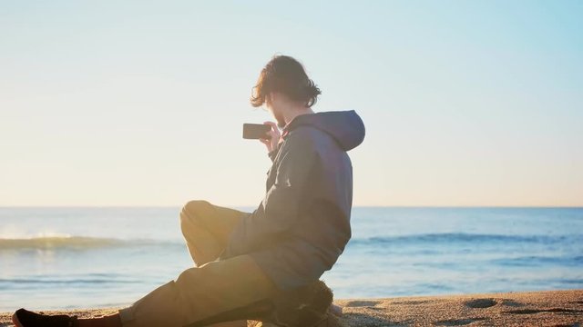 Young lonely nostalgic millennial man is having private melancholic moment and making picture or photo by smartphone camera in loneliness alone in beach watching sunrise or sunset sky and sea or ocean