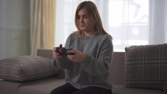 Portrait plus size girl playing video game sitting at sofa in home. Discontented chubby woman beats hand on broken joystick. Leisure of plus size lady at home indoors