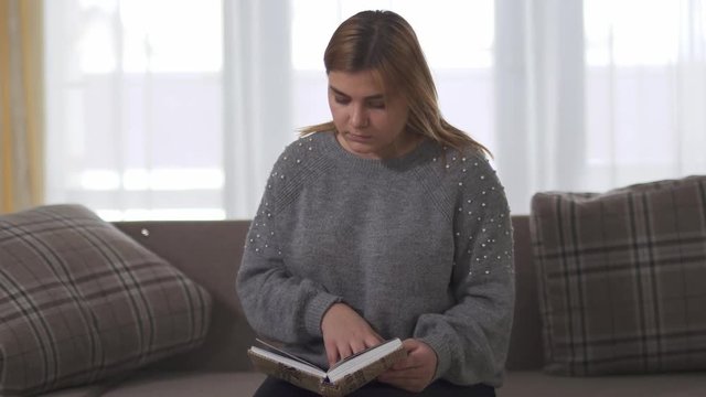 Plump girl reading book sitting at sofa in home. Chubby woman holding book in hands. Leisure of smart plus size lady at home