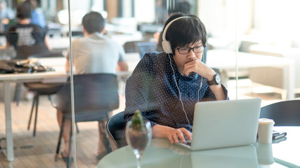 Young Asian business man with glasses and headphones working with laptop computer in coworking...
