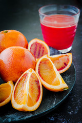 Fresh Juicy Blood Oranges Whole and Sliced on Dark Plate with Glass of Juice in Background
