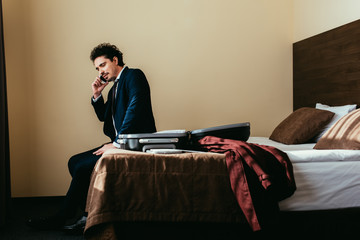 businessman talking on smartphone while sitting on bed with suitcase in hotel room
