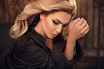 Gorgeous model. Blond woman portrait in black shirt. Fashionable girl with beauty makeup and curly hair style posing in wooden dark interior.